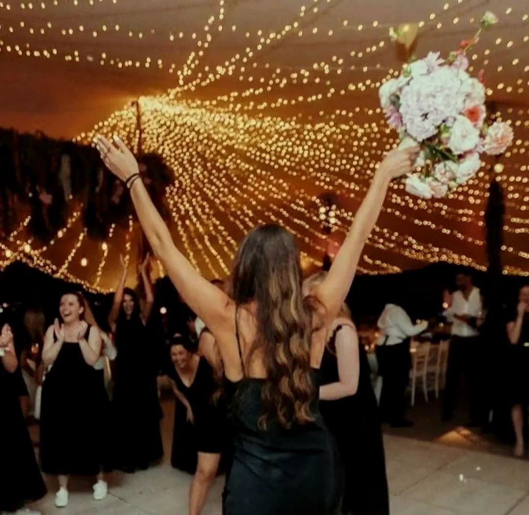 A guest with arms raised holding a bouquet on a dance floor, beneath a vast canopy of warm fairy lights