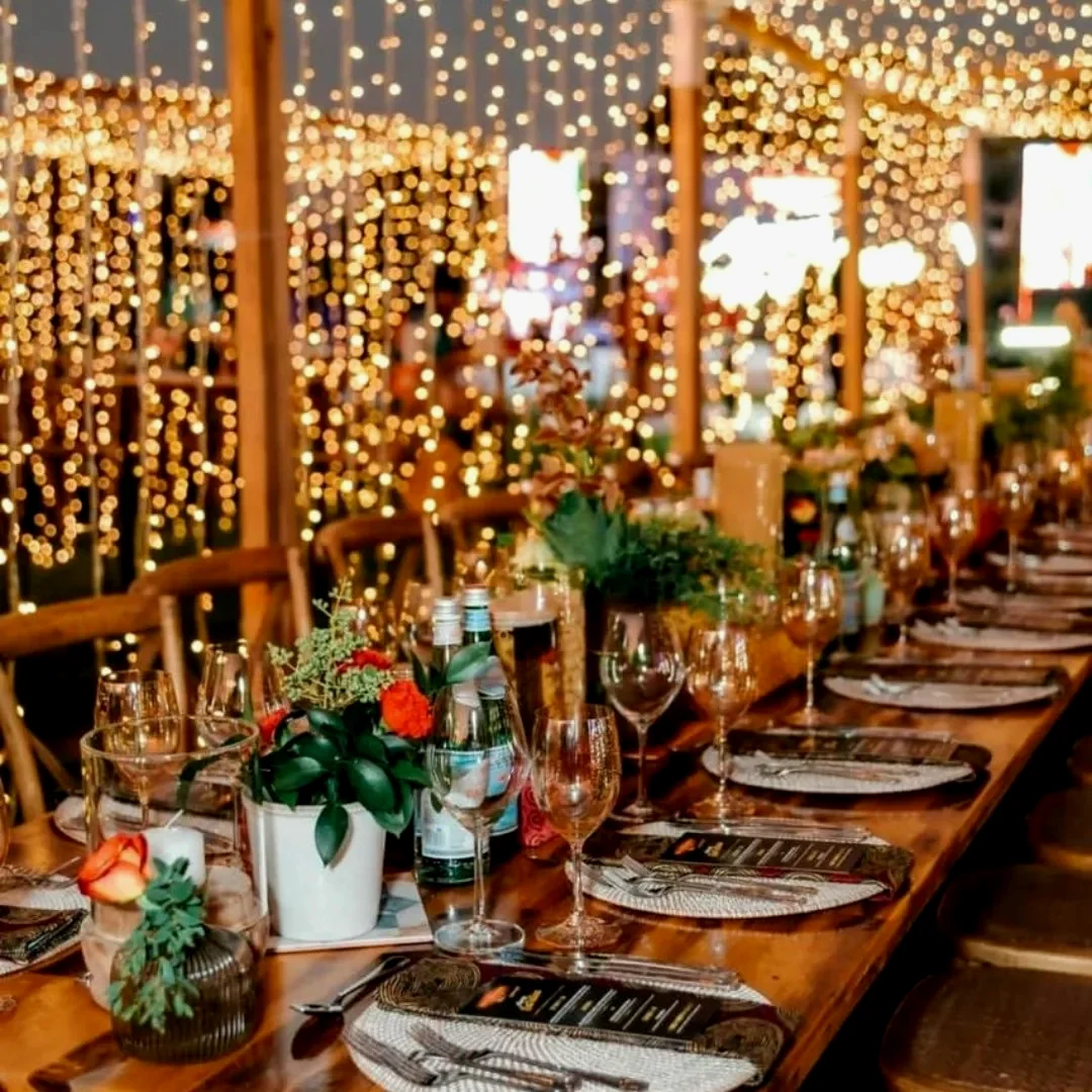 A long banquet table dressed with greenery and glassware, set in front of walls hung with curtains of fairy light