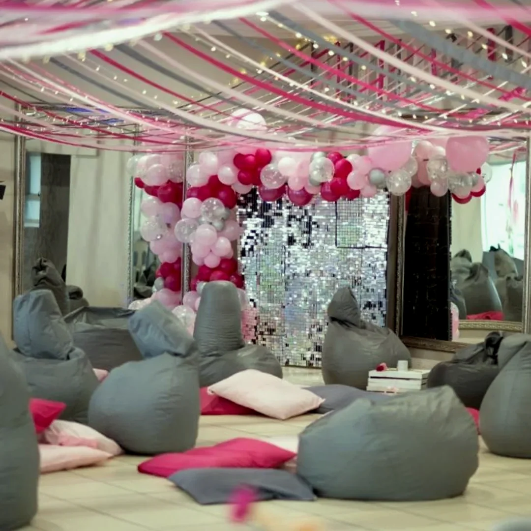 A function room with draped pink and white ceiling fabric, a balloon garland, a sequin backdrop and grey bean bags on the floor
