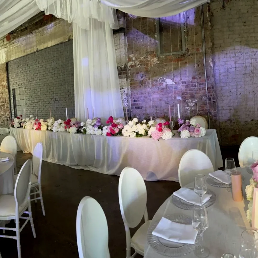 A long white-linen top table with pink and white florals and clear chairs, against a brick wall washed in lilac light beside pale draping