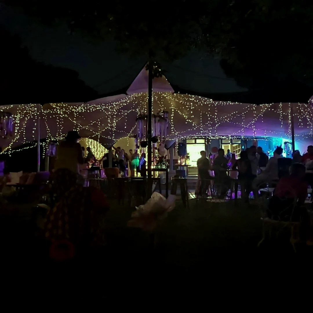 An outdoor evening function under a stretch tent, lit with warm fairy light and blue and purple wash, with guests in silhouette
