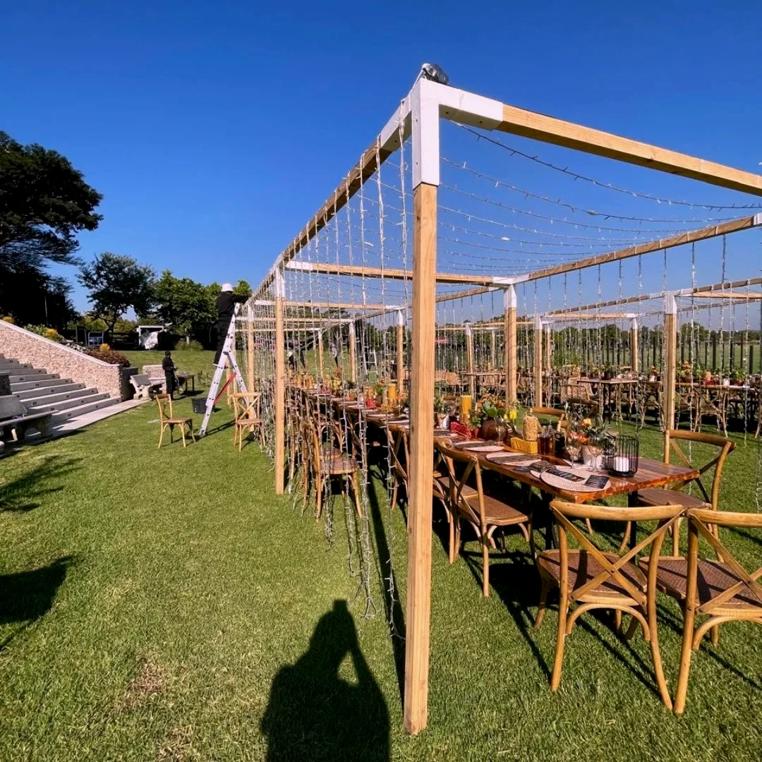 A timber walkway structure standing on a lawn in daylight, with tables and cane chairs set along it under a clear sky