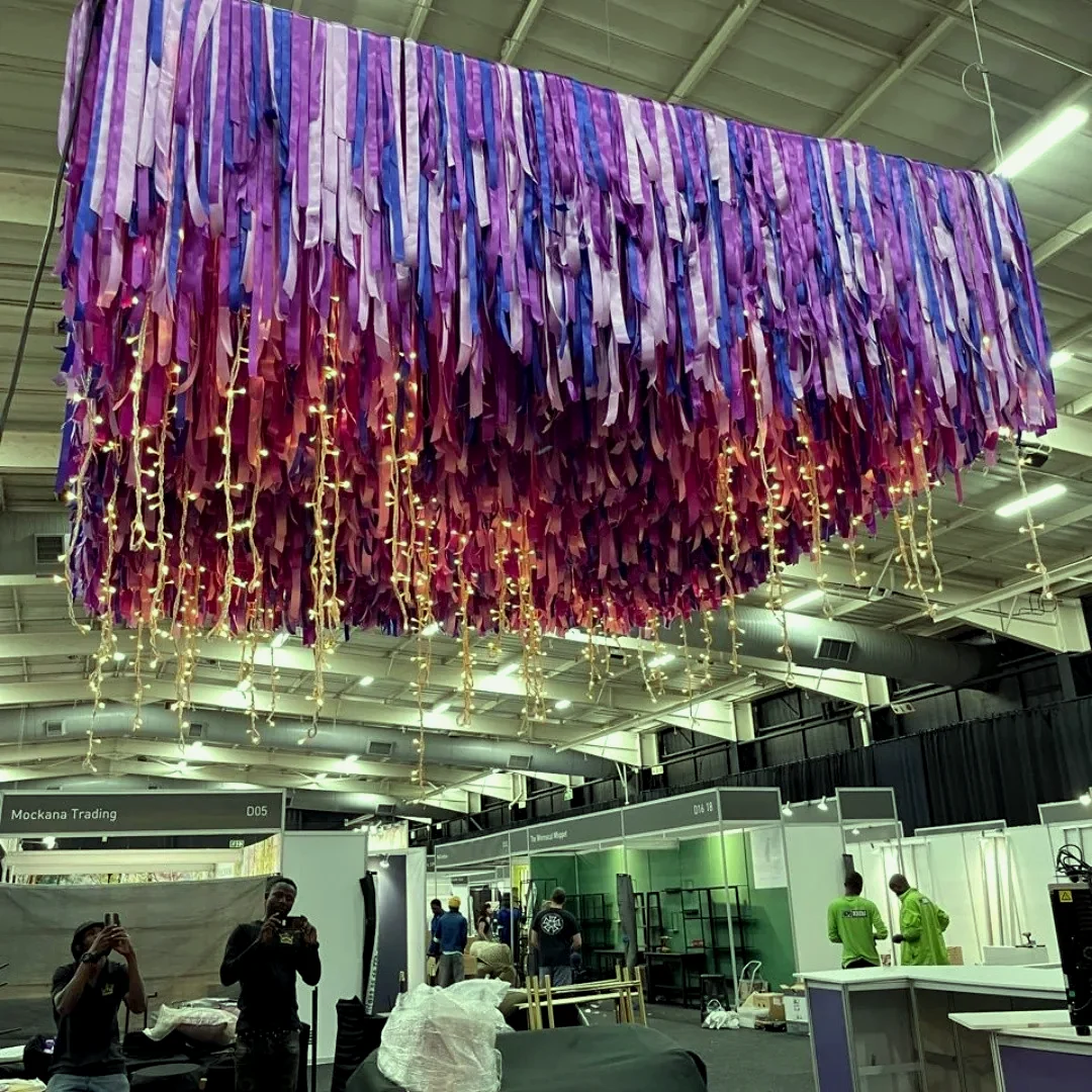 A wide hall with a suspended ceiling of purple and magenta fringe above an open exhibition floor, with people walking below